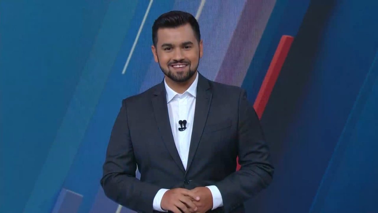 Yuri Macri, vestindo terno escuro e camisa branca aparece em estúdio de televisão, sorrindo para a câmera. O fundo tem tons de azul e vermelho, com o logotipo do jornal SBT Notícias