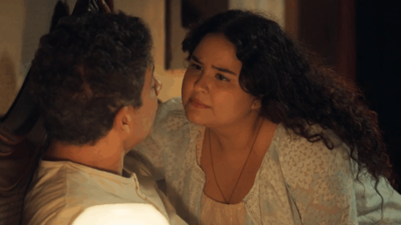 Cena mostra duas pessoas em um momento de proximidade e emoção. A mulher, com expressão séria e olhar intenso, está inclinada em direção ao homem que está sentado, sugerindo uma conversa importante ou tensa. O ambiente tem iluminação quente e acolhedora, típica de um cenário doméstico