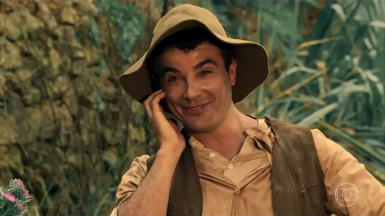 Homem com chapéu de palha sorri enquanto fala ao telefone em cenário rural em cena de novela