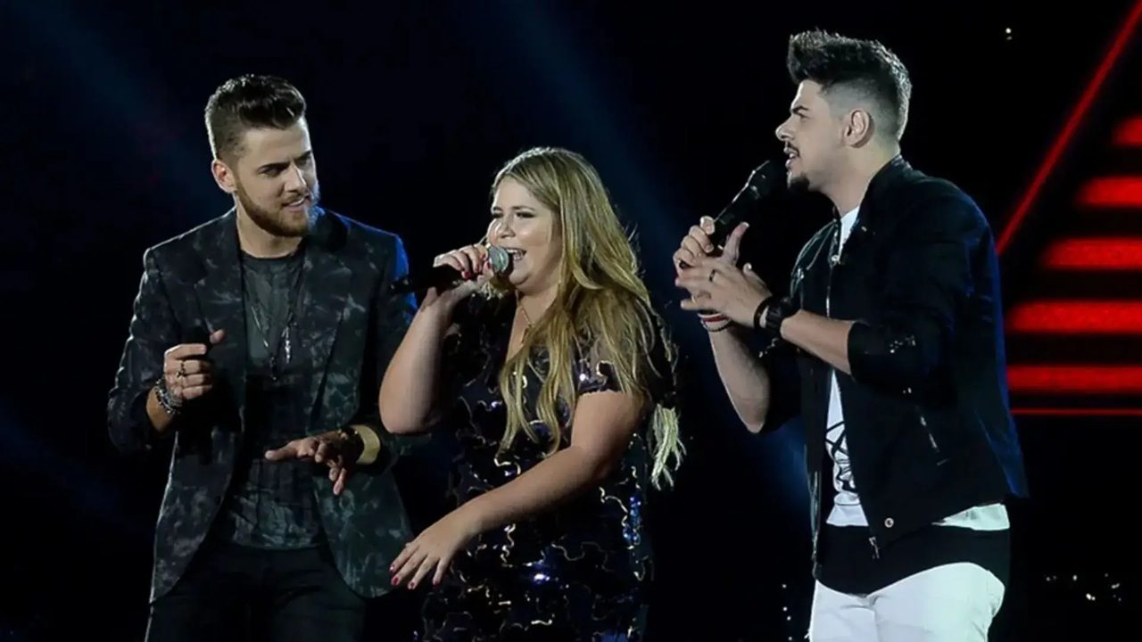 Três cantores se apresentam juntos em palco escuro iluminado por luzes de show com mulher ao centro cantando ao microfone usando vestido preto brilhante com cabelos longos soltos enquanto dois homens cantam ao lado dela cada um com microfone vestindo roupas escuras estilosas com fundo preto e detalhes de iluminação vermelha sugerindo apresentação musical ao vivo