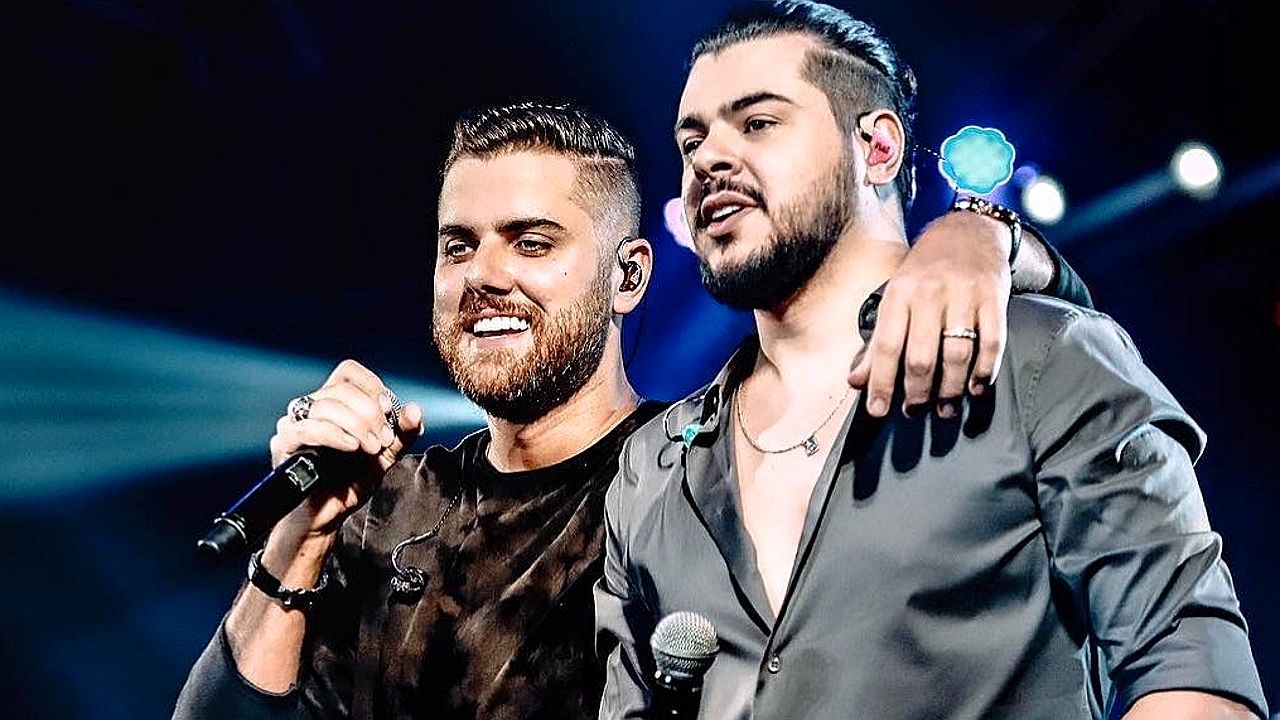 Dupla sertaneja canta abraçada no palco com microfones durante show ao vivo com luzes ao fundo
