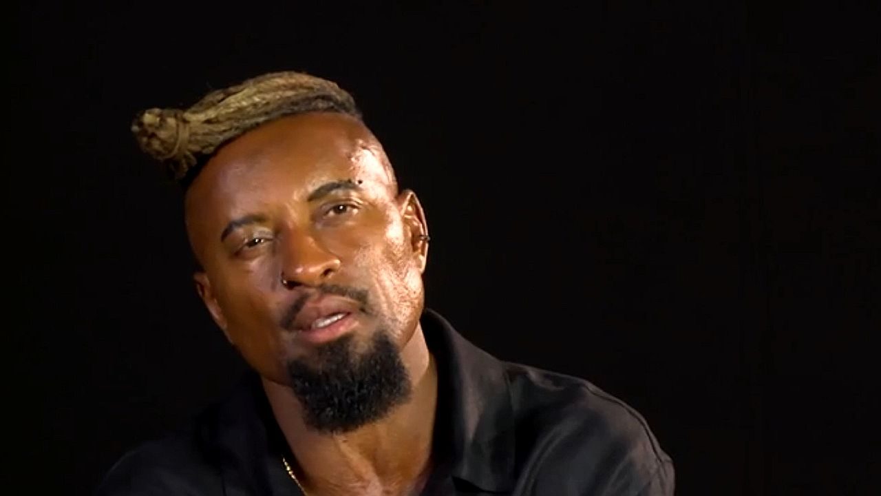 Homem com barba e cavanhaque, usando brinco e um penteado preso no topo da cabeça (tipo dread ou coque), falando ou sendo entrevistado diante de um fundo escuro.