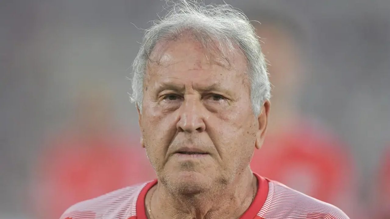 Homem de cabelos grisalhos veste camisa esportiva vermelha e branca e olha seriamente para frente em ambiente externo desfocado ao fundo