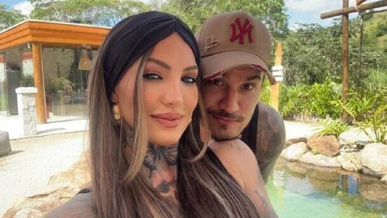 Mulher e homem posam juntos sorrindo levemente em frente a uma piscina ao ar livre, cercada por plantas e pedras. A mulher, com cabelos longos e lisos, usa uma faixa preta na cabeça e brincos dourados, enquanto o homem, de boné bege com logotipo vermelho, está atrás dela, com o braço apoiado em seu ombro. Ao fundo há uma construção moderna de madeira e vidro em meio a área arborizada.