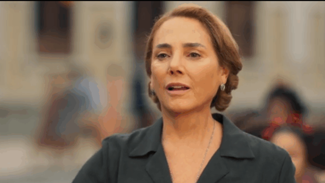 Mulher de expressão séria aparece em ambiente externo com construções históricas ao fundo, vestindo roupa escura e colar discreto, em cena de tom dramático